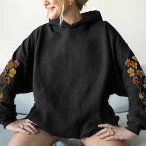 May include: Black hooded sweatshirt with floral embroidery on the sleeves. The flowers are in shades of orange, yellow, and pink, with green leaves. The sweatshirt has a front pocket and a drawstring hood.