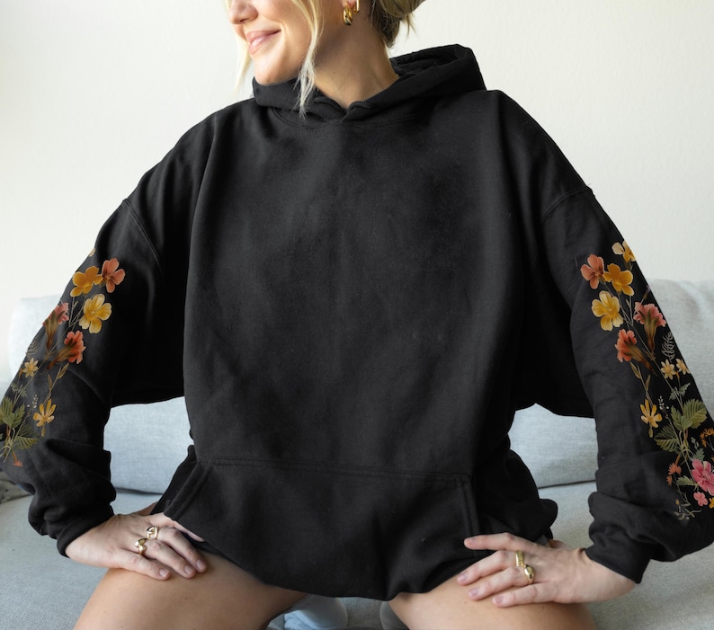 May include: Black hooded sweatshirt with floral embroidery on the sleeves. The flowers are in shades of orange, yellow, and pink, with green leaves. The sweatshirt has a front pocket and a drawstring hood.