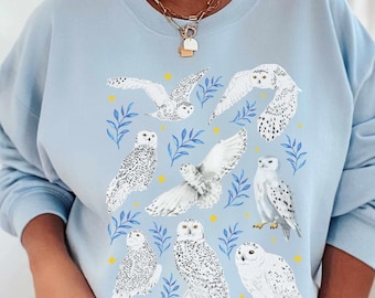 May include: A light blue crewneck sweatshirt featuring a graphic print of seven snowy owls in various poses, interspersed with small blue leaf sprigs and yellow four-pointed stars. The owls are depicted in white with black speckles.