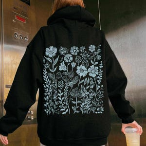 May include: Black hoodie with a light blue floral design on the back. The design features a variety of flowers and leaves.