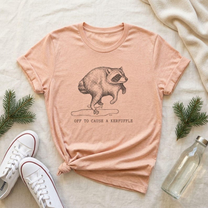 May include: A peach-colored short-sleeve t-shirt featuring a black line-art illustration of a raccoon stepping into a puddle. Below the raccoon, the text reads OFF TO CAUSE A KERFUFFLE in a simple, dark, typewriter-style font centered on the chest.