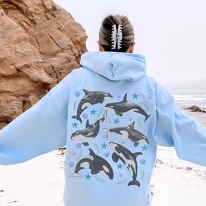May include: A light blue hoodie featuring a graphic print of six black and white orcas, seashells, and starfish. The orcas are swimming in various directions and are surrounded by seaweed.