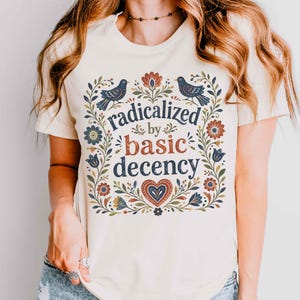 May include: A cream-coloured short-sleeve t-shirt featuring a central graphic with the text "radicalized by basic decency" in dark blue and rust orange. The design is surrounded by a floral wreath with two birds and a heart at the bottom.