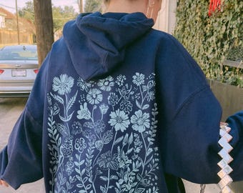 May include: A dark navy blue hooded sweatshirt featuring a large, light blue graphic print on the back. The design consists of various line-drawn wildflowers, stems, and leaves arranged in a dense, vertical botanical pattern across the fabric.