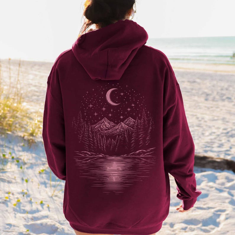 May include: Burgundy hoodie with a nature-themed graphic on the back. The design features a crescent moon, stars, mountains, trees, and a lake. The hoodie has a drawstring hood and a kangaroo pocket.