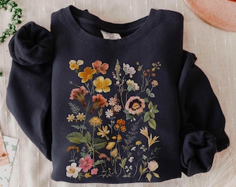 May include: A dark navy blue crewneck sweatshirt featuring a central botanical print of various wildflowers in shades of yellow, orange, pink, and white, with green foliage and stems arranged in a detailed, multi-colored floral illustration.