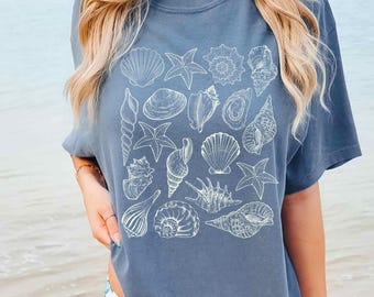 Op de afbeelding: Een leiblauw t-shirt met korte mouwen en een witte lijntekening-print van diverse schelpen en zeesterren in een groep op de voorzijde. Het ontwerp bevat gedetailleerde illustraties van verschillende soorten schelpen en zeesterren.