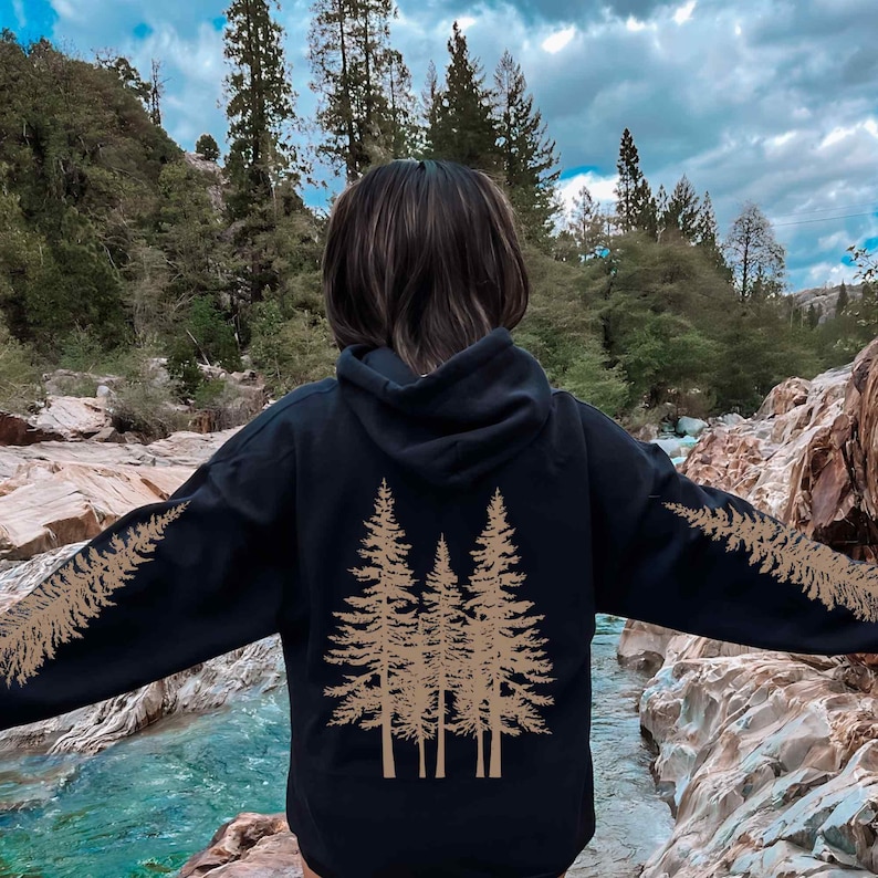 May include: Black hoodie with a nature-inspired design. The back features a cluster of three stylized pine trees in a tan color. Matching tan pine tree branches extend along the sleeves. The hoodie is made of a soft material and has a hood.