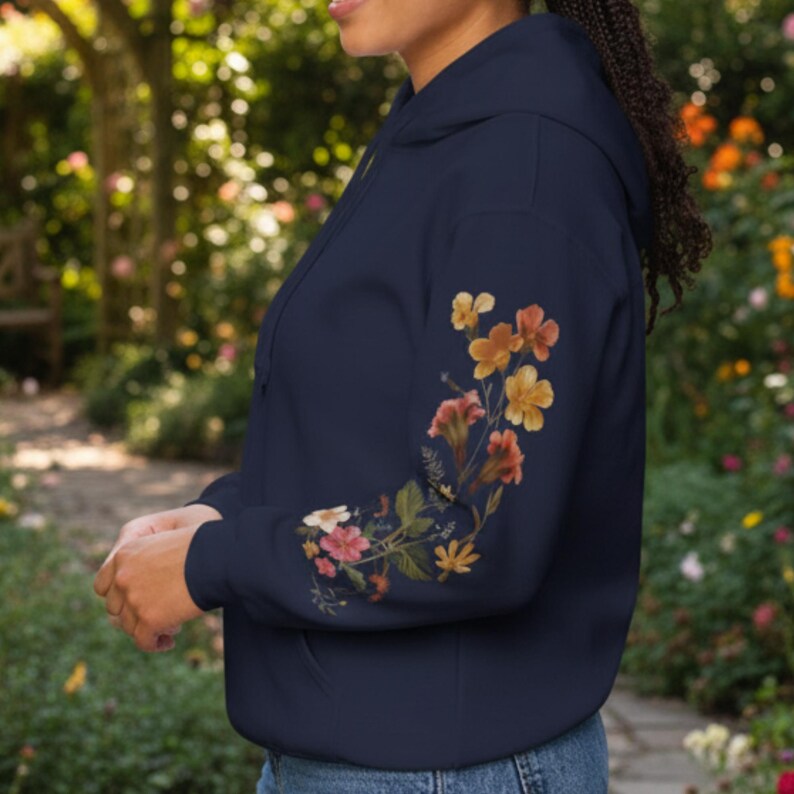 May include: Navy blue hoodie with a floral design on the sleeve. The flowers are in shades of orange, yellow, and pink, with green leaves. The hoodie has a drawstring hood and a front pocket.