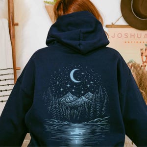 May include: Navy blue hoodie featuring a nature-themed graphic on the back. The design includes a crescent moon, stars, mountains, trees, and a lake. The hoodie has a hood and is made of a soft material.