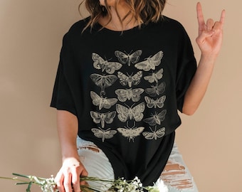 May include: A black short-sleeved t-shirt featuring a graphic print of various moth species in a light beige, line-art style. The design is arranged in a grid-like pattern across the center of the chest, showcasing detailed wing textures and shapes.