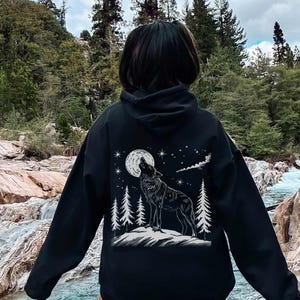 May include: Black hoodie featuring a white graphic of a howling wolf under a full moon and stars. The design includes stylized pine trees. The hoodie is worn outdoors near a river.