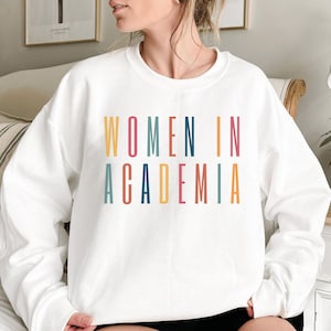Könnte beinhalten: Ein weißes Langarm-Sweatshirt mit Rundhalsausschnitt und dem Text "WOMEN IN ACADEMIA", der in einer vertikalen, mehrfarbigen serifenlosen Schrift gedruckt ist. Die Buchstaben sind in Gelb, Petrol, Dunkelblau, Orange und Pink gehalten.