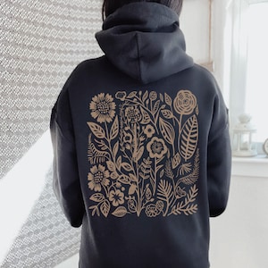 K&ouml;nnte beinhalten: Schwarzer Kapuzenpullover mit einem braunen Blumendruck auf der R&uuml;ckseite. Der Druck zeigt eine Vielzahl von Blumen, Bl&auml;ttern und Stielen.