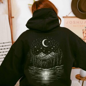 May include: Black hoodie featuring a nature-themed graphic on the back. The design includes a crescent moon, stars, mountains, trees, and a lake. The hoodie has a hood and a relaxed fit, suitable for casual wear.