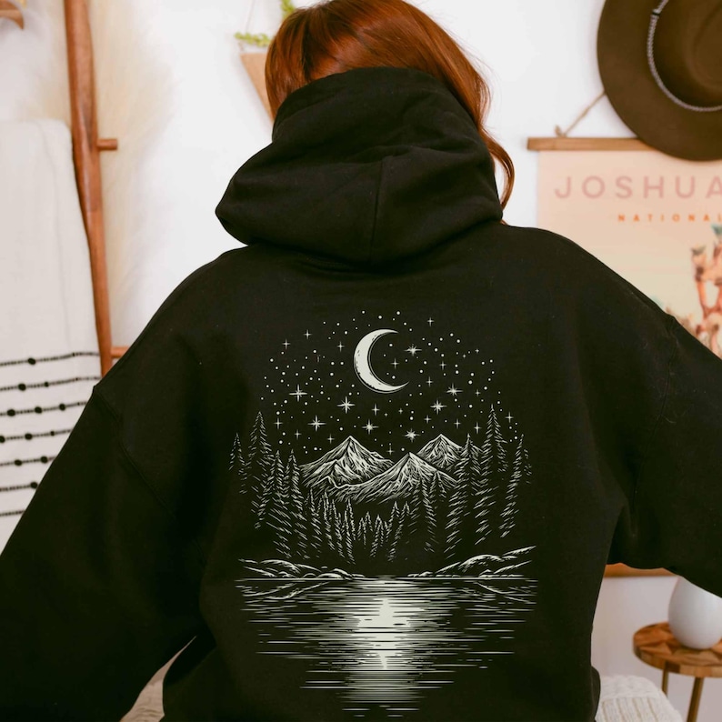 May include: Black hoodie featuring a nature-themed graphic on the back. The design includes a crescent moon, stars, mountains, trees, and a lake. The hoodie has a hood and a relaxed fit, suitable for casual wear.