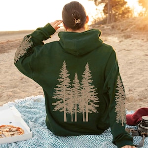 May include: A dark green hoodie with a tan tree graphic on the back and sleeves. The hoodie is oversized and has a hood. The person is sitting on a blanket with a pizza box and camera nearby. The hoodie is made of a soft material.