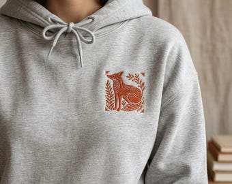 May include: A light gray hooded sweatshirt featuring a drawstring hood and a square embroidered patch on the chest. The patch displays a rust-colored fox sitting among stylized leaves and branches, rendered in a detailed, linear embroidery style.