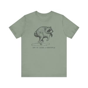 May include: A sage green short-sleeve t-shirt featuring a black ink-style illustration of a raccoon walking on its hind legs. Below the raccoon is a small puddle and the text "OFF TO CAUSE A KERFUFFLE" printed in a simple, dark typewriter-style font.