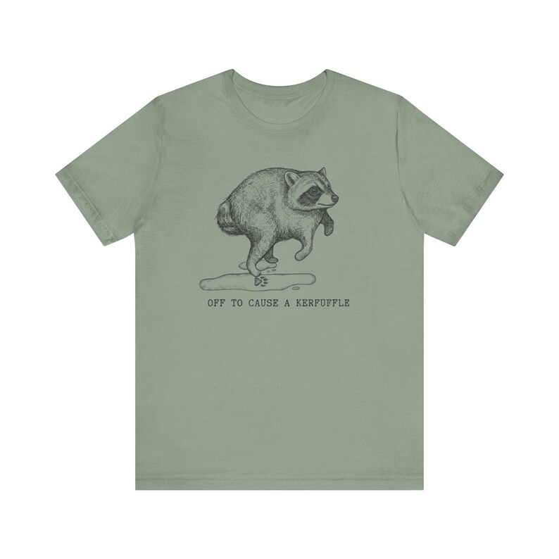 May include: A sage green short-sleeve t-shirt featuring a black ink-style illustration of a raccoon walking on its hind legs. Below the raccoon is a small puddle and the text "OFF TO CAUSE A KERFUFFLE" printed in a simple, dark typewriter-style font.