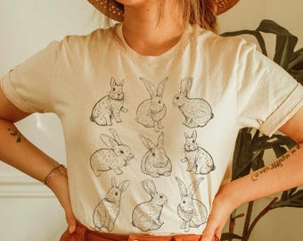 May include: A cream-colored short-sleeve t-shirt featuring a grid of nine black line-art illustrations of rabbits in various poses. The rabbits are depicted in different positions, including sitting, crouching, and facing forward, arranged in three rows.