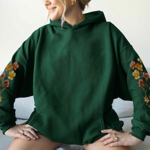 May include: A dark green hoodie with a front pocket and a hood. The sleeves are decorated with floral embroidery in shades of orange, yellow, and pink. The hoodie is made of a soft material.