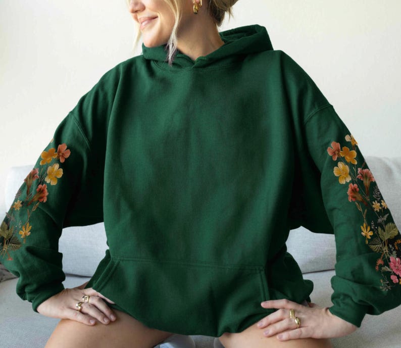 May include: A dark green hoodie with a front pocket and a hood. The sleeves are decorated with floral embroidery in shades of orange, yellow, and pink. The hoodie is made of a soft material.