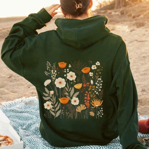 Puede incluir: Una sudadera con capucha verde oscuro con un estampado floral en la espalda. El estampado presenta una variedad de flores en tonos naranja, amarillo y blanco.