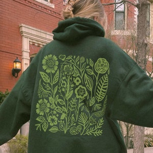 Könnte beinhalten: Ein dunkelgrüner Hoodie mit einem großen grünen Blumendruck auf der Rückseite. Der Druck zeigt stilisierte Blumen, Blätter und Stängel.