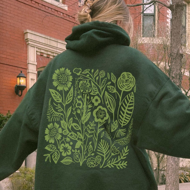 May include: A dark green hoodie with a large green floral print on the back. The print features stylized flowers, leaves, and stems.