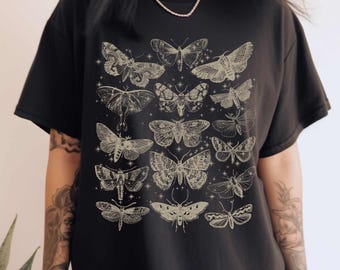 May include: A black short-sleeved t-shirt featuring a graphic print of various moth species arranged in a grid pattern. The moths are illustrated in a light beige color with intricate wing details and small star accents scattered throughout the design.