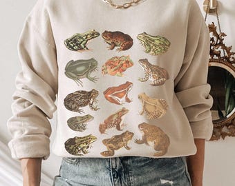 May include: A beige crewneck sweatshirt featuring a graphic print of twelve different frogs and toads arranged in a grid. The illustrations display various species in shades of green, brown, and orange, showcasing distinct textures and markings.