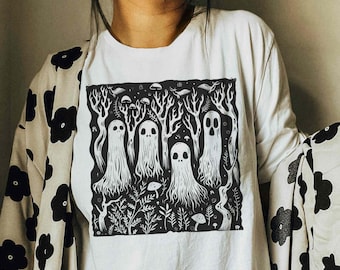 May include: A white long-sleeve t-shirt featuring a black square graphic print of four ghosts standing in a forest with trees, ferns, and mushrooms. The shirt is worn with a light-colored cardigan patterned with large black flower shapes.