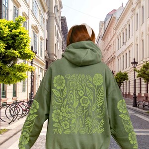 Puede incluir: Sudadera con capucha verde con un dise&ntilde;o floral en verde claro en la espalda y las mangas. El dise&ntilde;o presenta varias flores y hojas. La sudadera tiene capucha y un corte holgado, adecuada para uso casual.