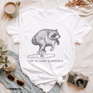 May include: A white short-sleeved t-shirt featuring a black and white line art illustration of a raccoon stepping into a puddle. Below the raccoon, the text reads OFF TO CAUSE A KERFUFFLE in a simple, dark, sans-serif font style.