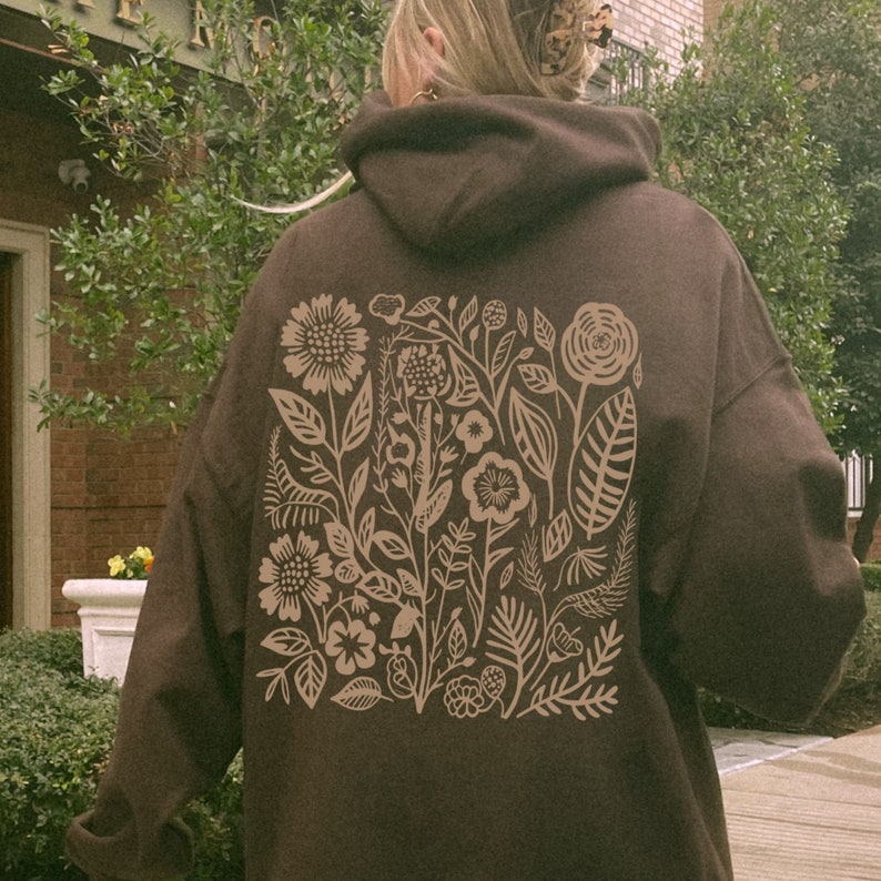 K&ouml;nnte beinhalten: Ein brauner Hoodie mit einem floralen Design, das auf der R&uuml;ckseite aufgedruckt ist. Das Design zeigt eine Vielzahl von Blumen, Bl&auml;ttern und Stielen in einer hellbraunen Farbe.