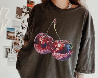 May include: A charcoal grey short-sleeve t-shirt featuring a graphic print of two cherries. The cherries are designed to look like reflective disco balls with a multi-colored, mirrored mosaic pattern in shades of pink, blue, and purple.