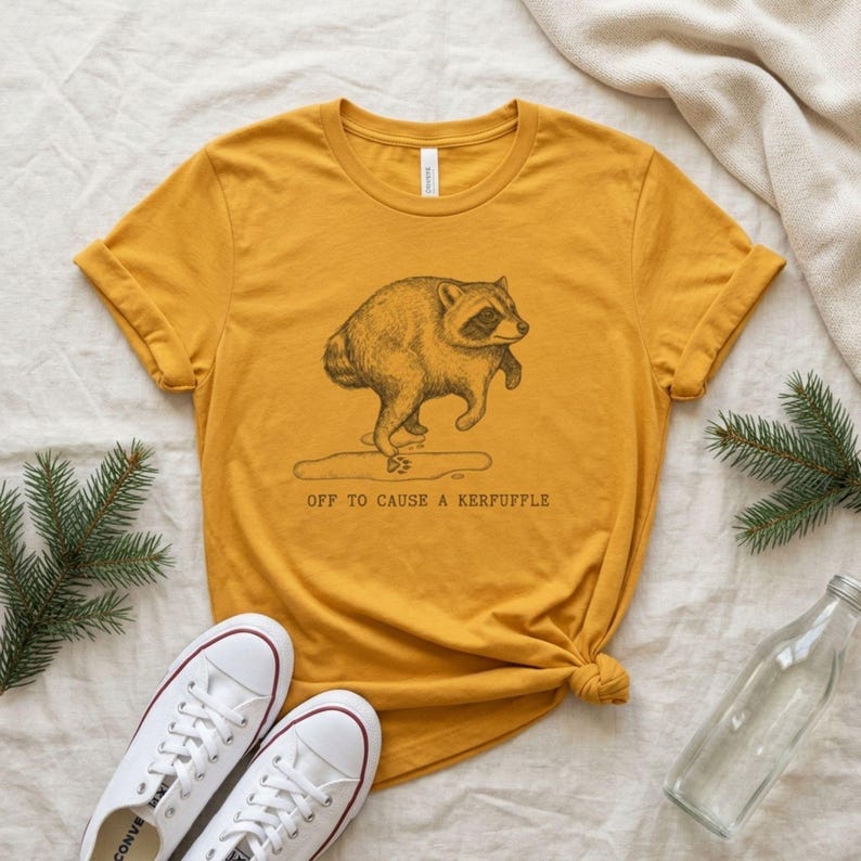 May include: A mustard yellow t-shirt featuring a black line art illustration of a raccoon standing on its hind legs. Below the raccoon is a small puddle and the text "OFF TO CAUSE A KERFUFFLE" printed in a simple, dark, sans-serif font.
