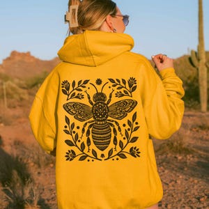 May include: A mustard yellow hooded sweatshirt featuring a large black graphic on the back. The design depicts a detailed bee surrounded by symmetrical floral and leaf patterns. The hoodie has long sleeves and a hood, showcasing a bold contrast.