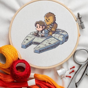 May include: A cross-stitch embroidery hoop featuring a cartoon illustration of Han Solo and Chewbacca in the Millennium Falcon. The image is surrounded by embroidery floss in shades of yellow, orange, and red, along with scissors, a thimble, and pins.