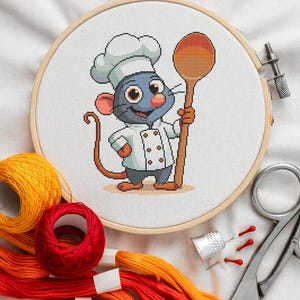 May include: A blue cartoon mouse wearing a white chef's hat and holding a wooden spoon. The mouse is embroidered on a white fabric stretched over a wooden embroidery hoop.