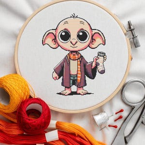 Może przedstawiać: Obręcz do haftu krzyżykowego z kolorowym obrazkiem Dobby'ego, domowego elfa z kreskówek, trzymającego skarpetkę. Haft wykonany jest na białym materiale, a obręcz jest z naturalnego drewna.