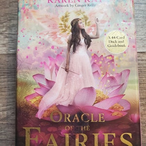 Peut inclure: Un livre intitulé "Oracle des Fées" de Karen Kay avec des illustrations de Ginger Kelly. La couverture présente une fée vêtue d'une robe rose debout sur un lotus rose. Le livre comprend un jeu de 44 cartes et un guide.