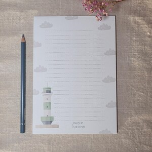 Könnte beinhalten: Ein liniertes Notizbuch mit einer Leuchtturm-Illustration und Wolkenmotiven. Der Leuchtturm ist weiß und grün, mit den Worten "moin hanne" unten. Ein blauer Bleistift liegt links neben dem Notizbuch.