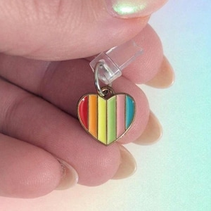 Puede incluir: Un pequeño dije en forma de corazón con un diseño de rayas de arcoíris. El dije tiene un borde dorado y un pequeño lazo de metal para sujetar. Las rayas del arcoíris incluyen rojo, naranja, amarillo, verde, rosa y azul.