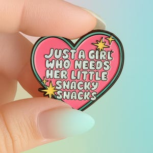 Op de afbeelding: Een hartvormige emaille speld met een roze centrum en een groenblauwe rand. De speld bevat de witte tekst "JUST A GIRL WHO NEEDS HER LITTLE SNACKY SNACKS" in een bubbellettertype, geaccentueerd door kleine gele vierpuntige sterren aan de zijkanten.