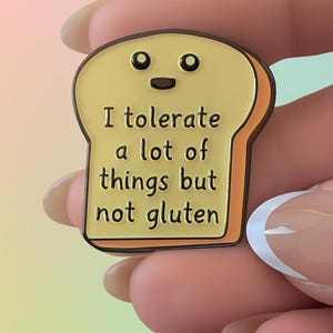 Puede incluir: Pin de esmalte con forma de rebanada de pan amarillo con una cara sonriente y el texto "I tolerate a lot of things but not gluten". El pin tiene un borde marrón y está sujeto por una mano. La marca "Anxious Oakley" está en la parte inferior.
