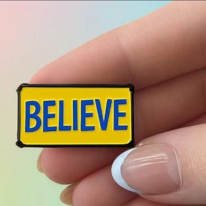 May include: A rectangular enamel pin with the word "BELIEVE" in blue letters on a yellow background, framed by a black border. The pin is held between fingers.