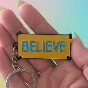 Peut inclure: Un porte-clés jaune avec le mot "BELIEVE" en lettres bleues. Le porte-clés a un porte-clés et une chaîne argentés. Le fond est un dégradé de rose, de bleu et de vert.