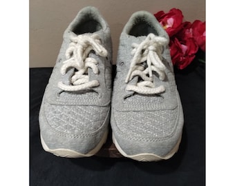 May include: A pair of gray sneakers with white laces. The shoes have a quilted pattern and a white sole. The sneakers are displayed on a black surface with red flowers in the background.